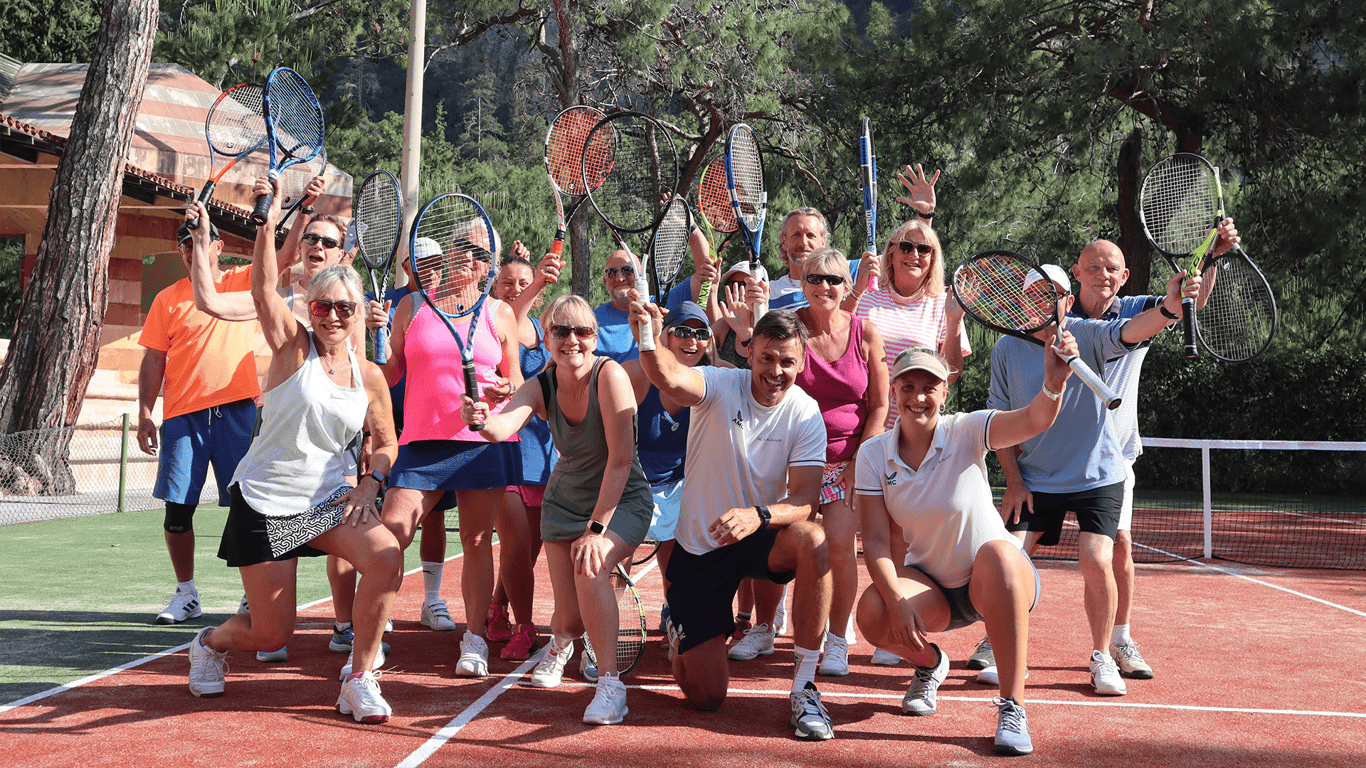 Smiling group of adults raising tennis racquets on an outdoor court during a holiday coaching session.