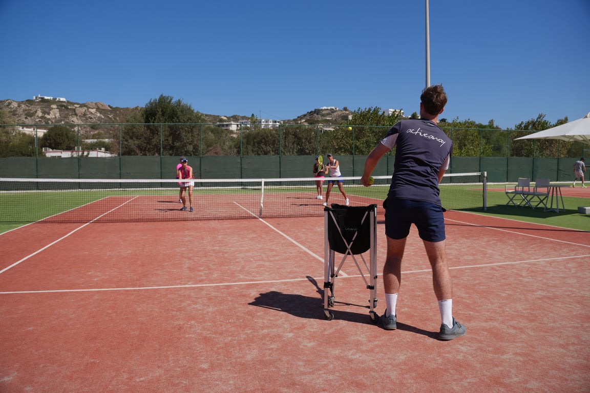 Tennis coach feeds balls to players at the net during a sunny coaching session on an outdoor clay court.