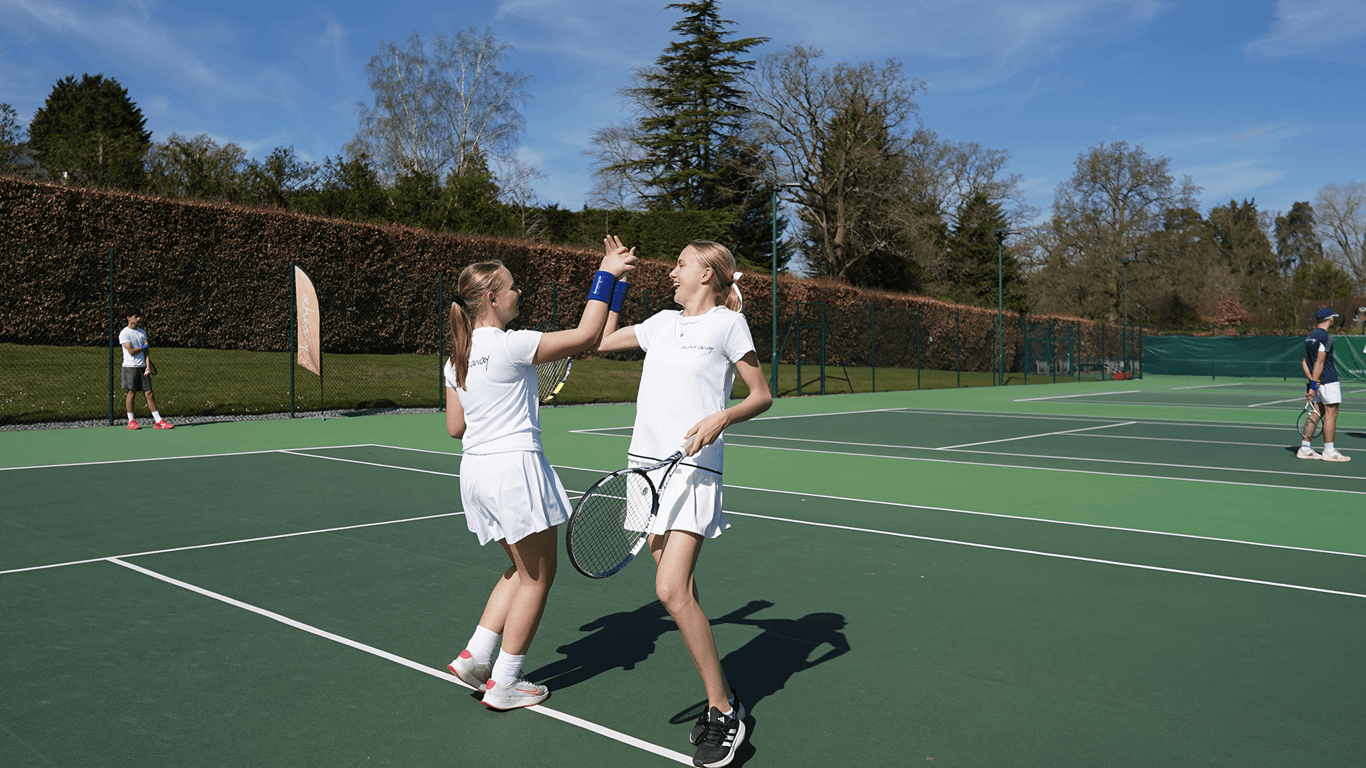 Two junior tennis players high-five on an outdoor court during practice, racquets in hand.