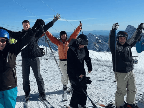 Group of skiers on a snowy mountain, smiling with arms raised under clear blue sky.