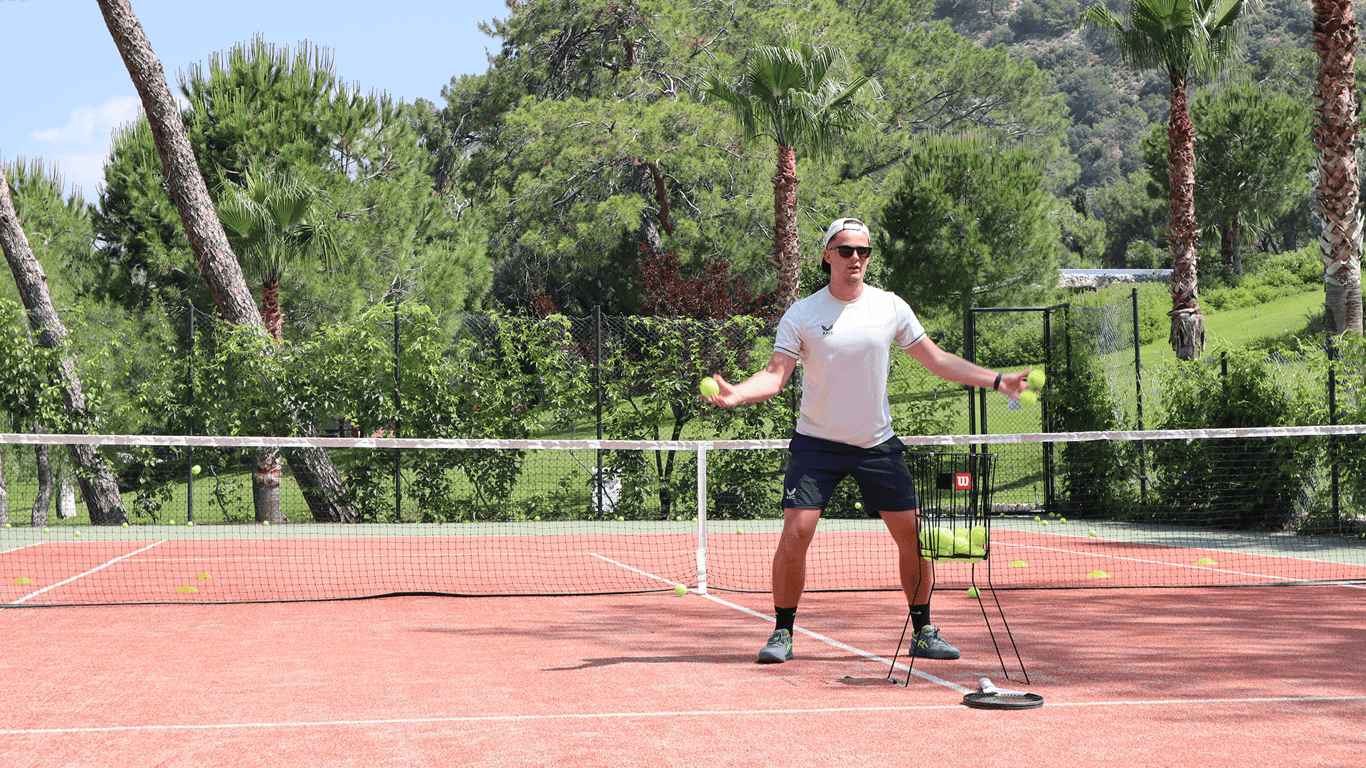Tennis coach demonstrating forehand drill on a sunny clay court with a basket of balls