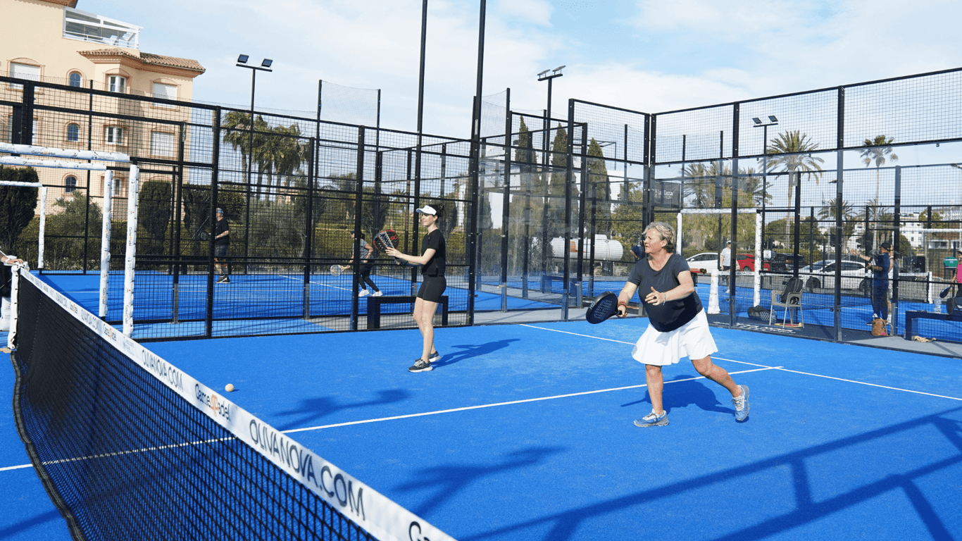 Active Away coach demonstrating a padel forehand on a blue outdoor court with players nearby.