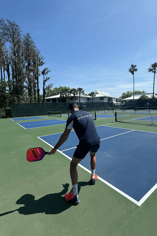 Man preparing to hit a pickleball on outdoor courts under a sunny sky, wearing an Active Away shirt.
