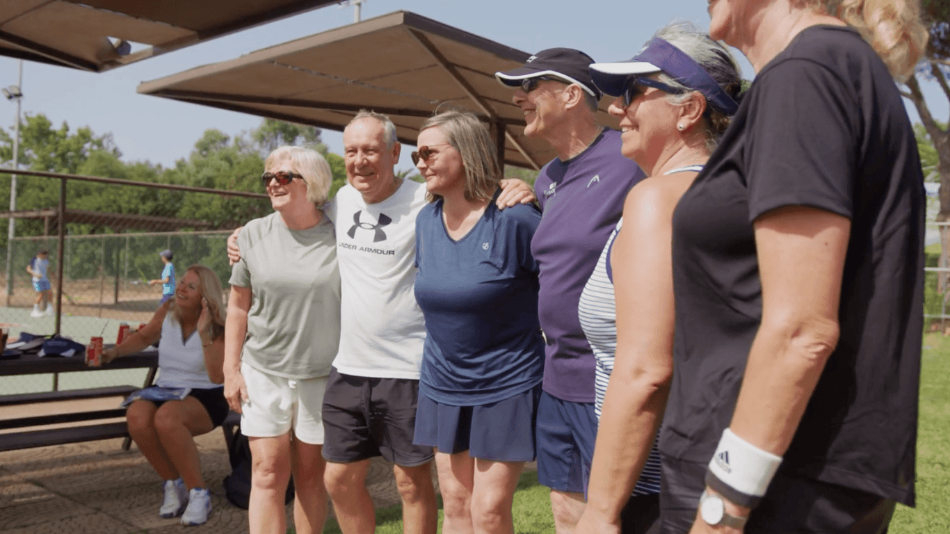 Smiling adults pose together beside outdoor courts on a pickleball holiday.