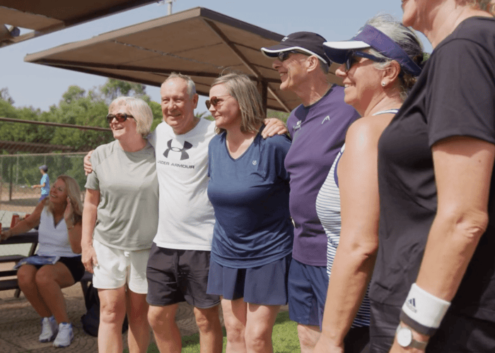 Smiling adults pose together beside outdoor courts on a pickleball holiday.