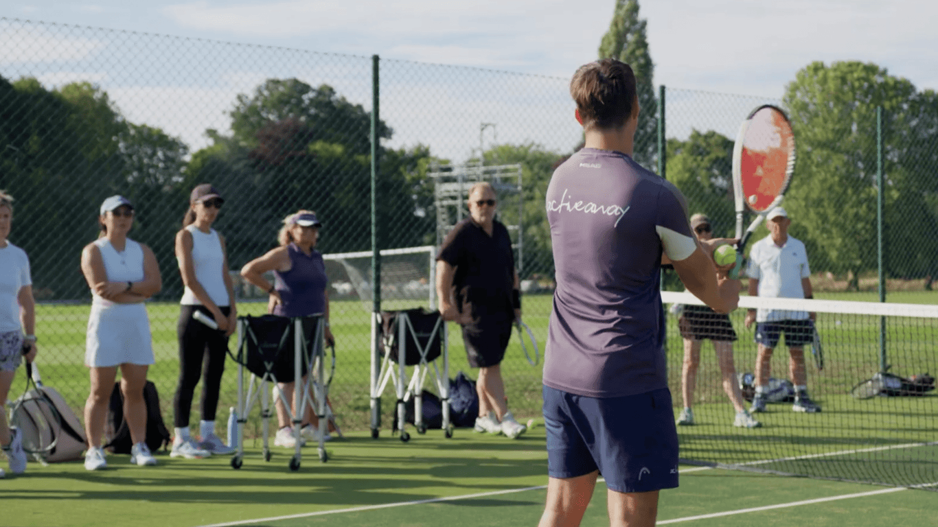 Active Away coach demonstrates forehand to adults during group lesson on Lensbury tennis courts.