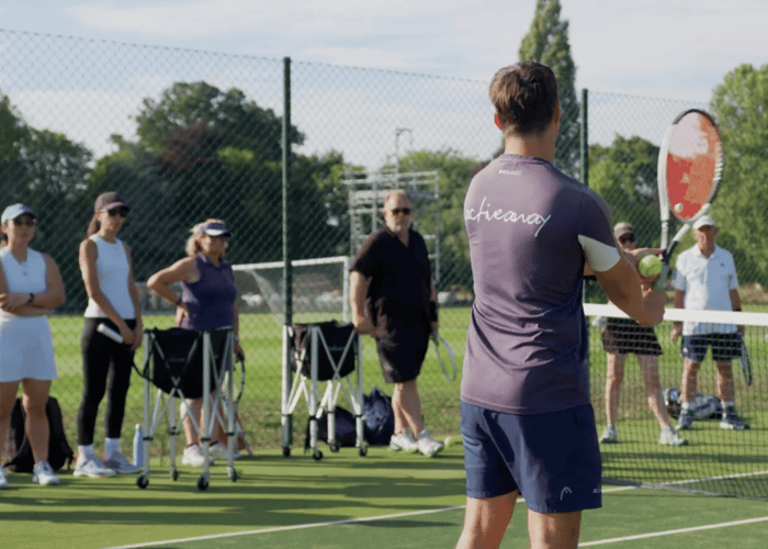Active Away coach demonstrates forehand to adults during group lesson on Lensbury tennis courts.