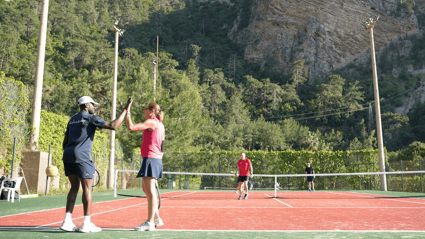 Two tennis players high-five on an outdoor red court with mountains and trees in the background.