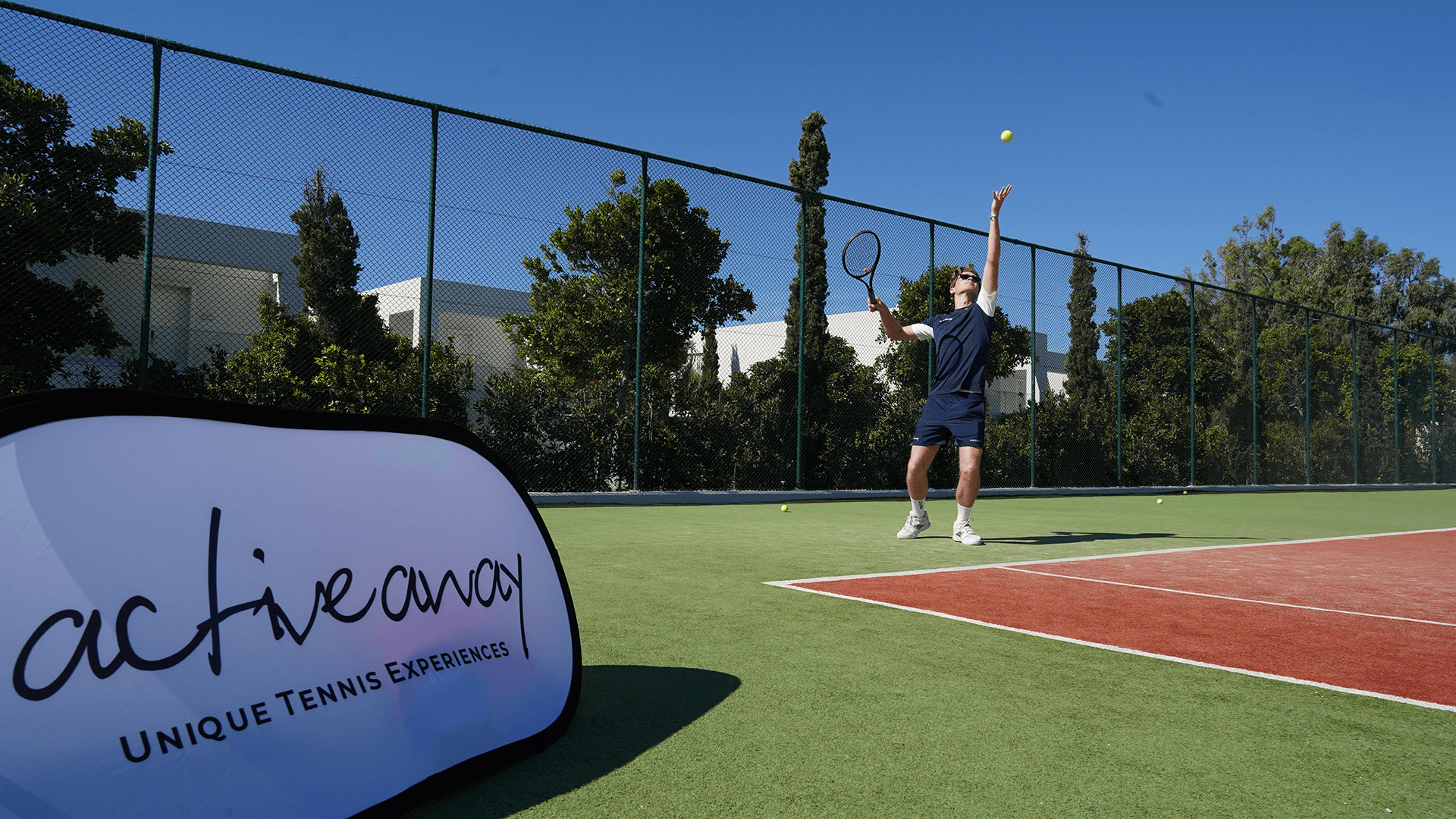 Tennis player serving on an outdoor court beside an Active Away sign.