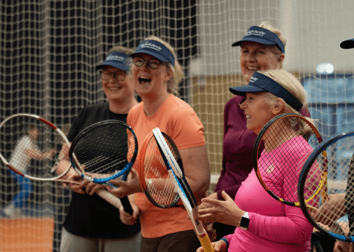 Group of smiling women holding tennis rackets at an indoor clinic, standing courtside in visors.