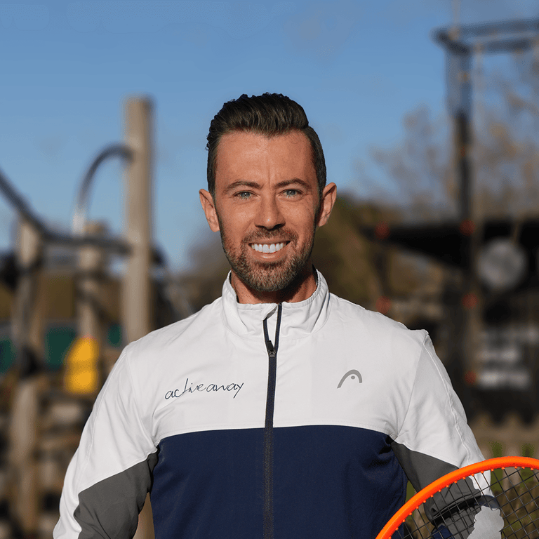 Portrait of a tennis coach in an Active Away jacket holding a racket on court.