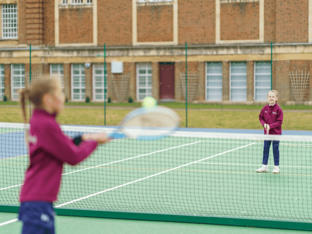 Children practising tennis on an outdoor court; foreground player hits a ball as partner waits across the net.