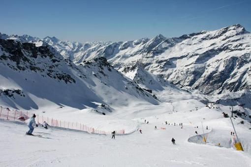 Skiers on a groomed slope at Alagna with snow-covered Alpine peaks under a clear sky.