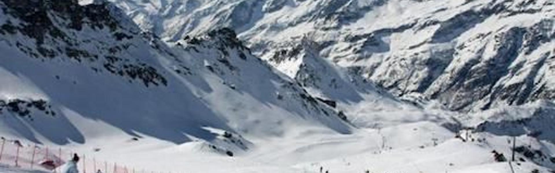 Skiers on a groomed slope at Alagna with snow-covered Alpine peaks under a clear sky.