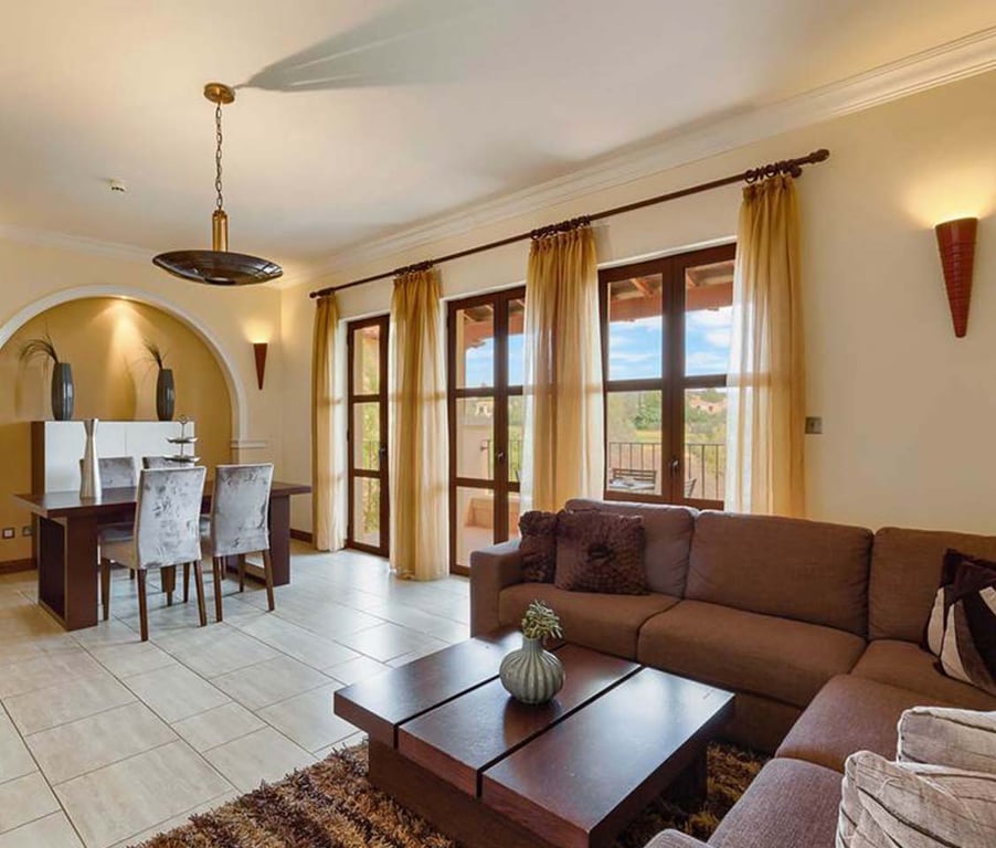 Living room of a one-bedroom apartment for six, with a brown sofa, wooden coffee table, and dining table for six, brightened by large windows.