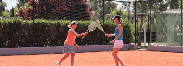 Two women in sportswear exchanging a racquet tap on a clay tennis court at Sani Resort.