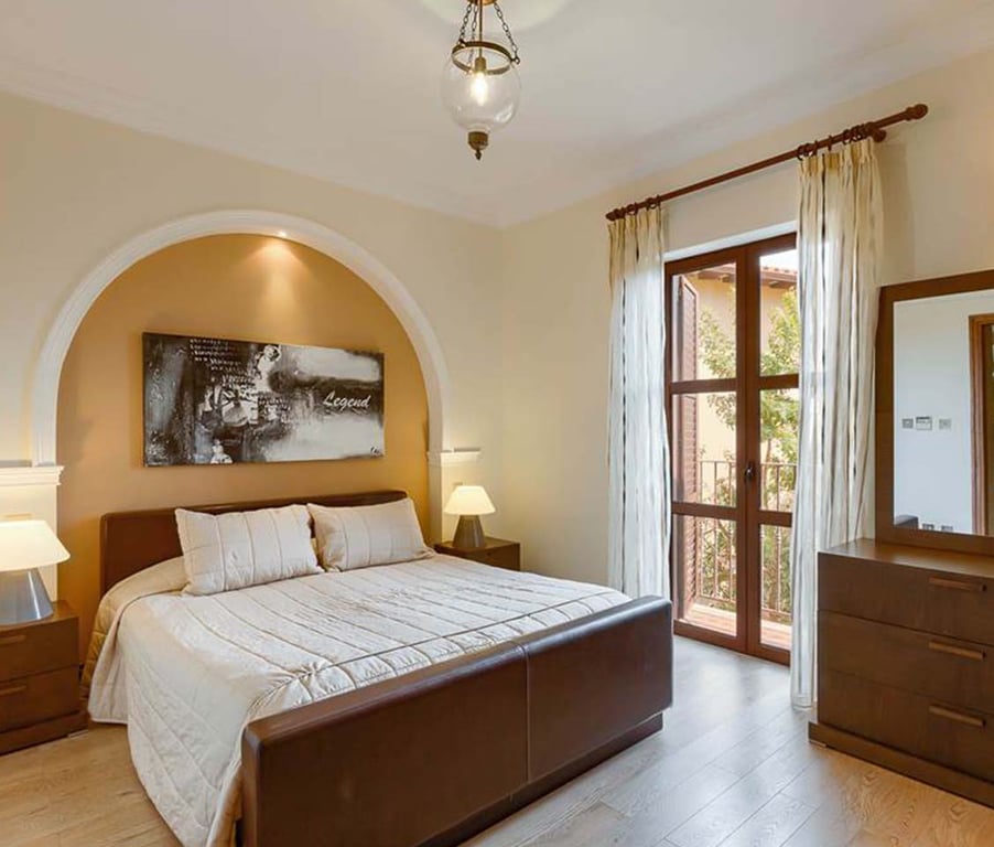 Bright master bedroom in a two-bedroom apartment, featuring a king-sized bed, beige decor, bedside tables, a window with curtains, and a dresser with a mirror.
