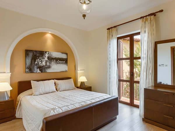 Bright master bedroom in a two-bedroom apartment, featuring a king-sized bed, beige decor, bedside tables, a window with curtains, and a dresser with a mirror.