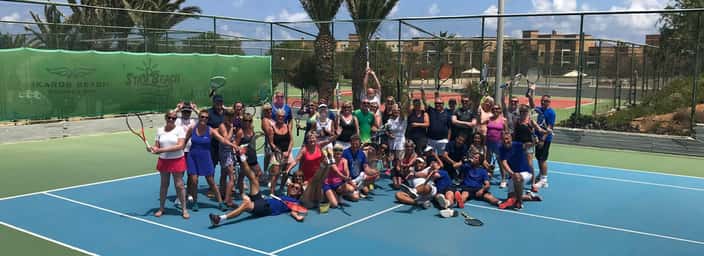 A group of people holding tennis rackets on an outdoor court, celebrating a 2018 tennis event under sunny skies.