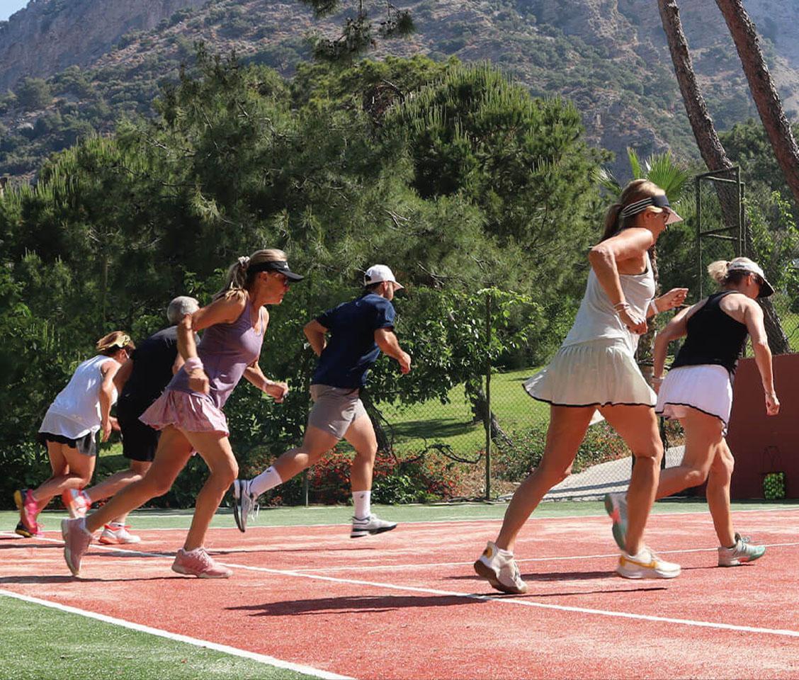 Athletes participate in fitness training on a tennis court amidst green foliage and mountains, highlighting the integration of tennis and fitness workouts.