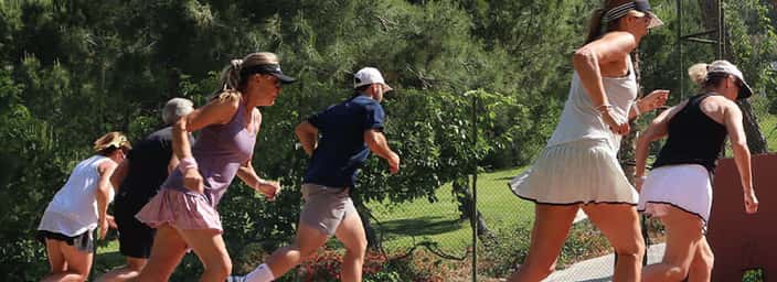 Athletes participate in fitness training on a tennis court amidst green foliage and mountains, highlighting the integration of tennis and fitness workouts.