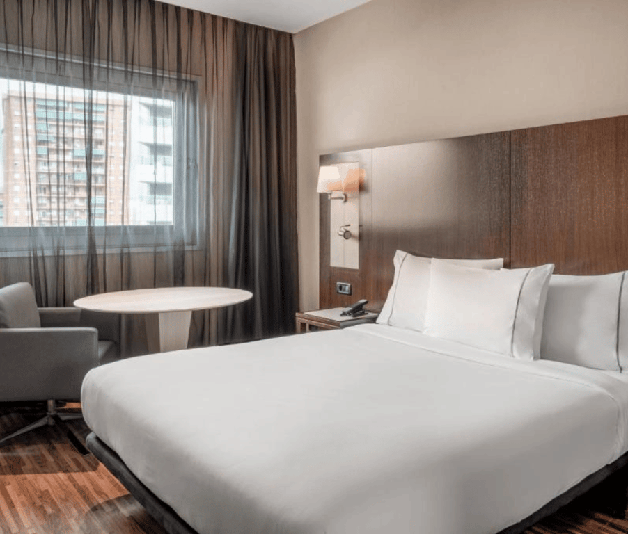 Standard room at AC Hotel with a queen-sized bed, wooden headboard, armchair, round table, and view of urban buildings through the window.