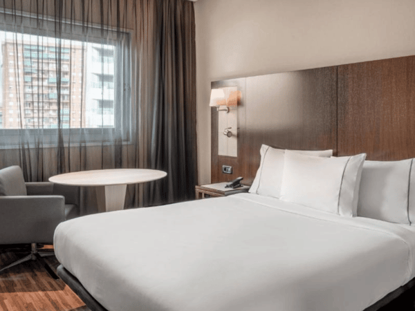 Standard room at AC Hotel with a queen-sized bed, wooden headboard, armchair, round table, and view of urban buildings through the window.