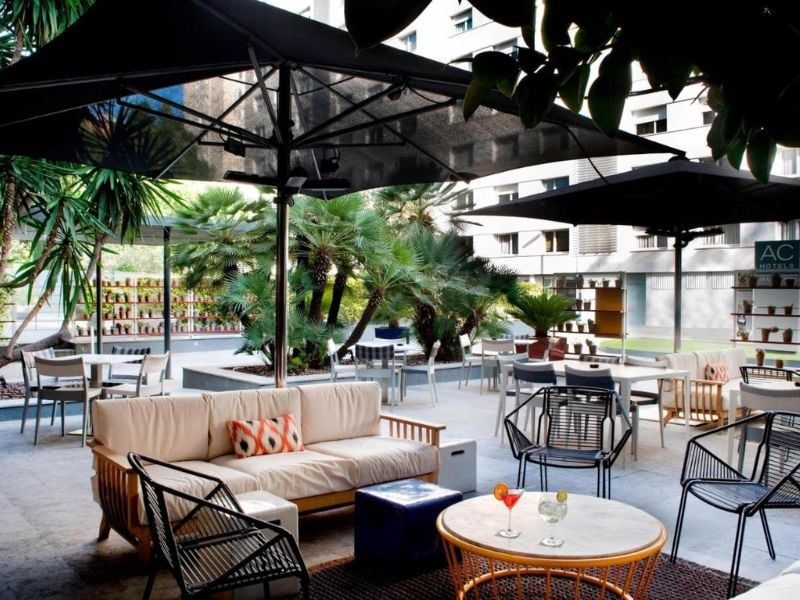 Outdoor dining area at AC Hotels with modern furniture, large umbrellas, palm trees, and a relaxed atmosphere.