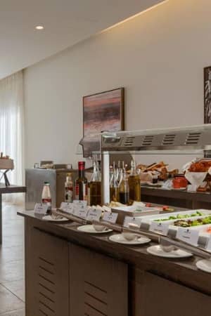 Buffet spread at Aphrodite Hills with fresh vegetables, salads, bread, condiments, oils, and modern decor in the background.