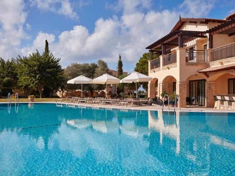Luxury villa at Aphrodite Hills Resort with a pool, sun loungers, and white umbrellas under a clear blue sky.