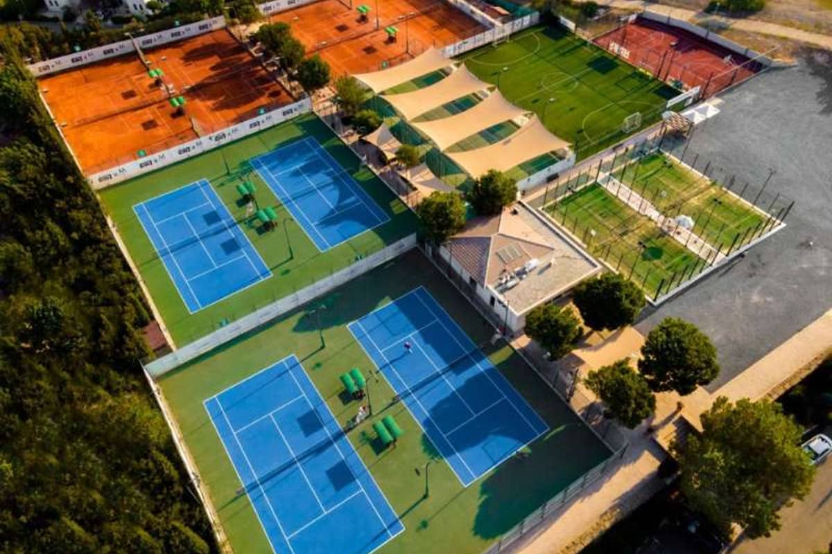 Aerial view of Aphrodite Hills Resort tennis courts, including blue hard courts and orange clay courts, surrounded by green fields and trees.