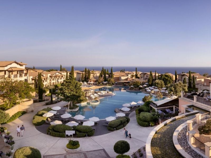 Aerial view of Aphrodite Hills Resort with a large pool, sun loungers, Mediterranean-style buildings, and a view of the sea.