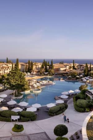 Aerial view of Aphrodite Hills Resort with a large pool, sun loungers, Mediterranean-style buildings, and a view of the sea.