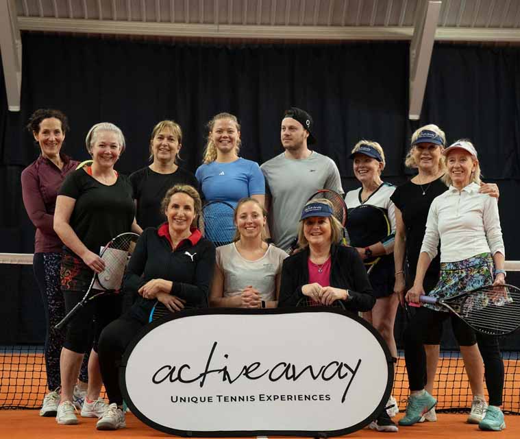 Group of adult tennis players at an indoor clinic, posing with an Active Away sign on a tennis court.