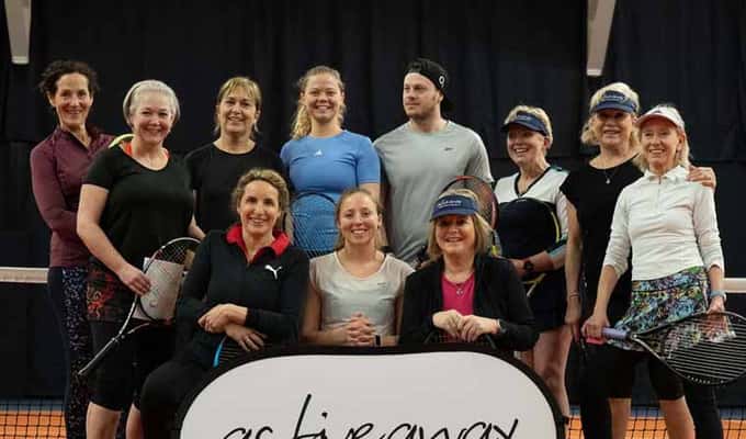 Group of adult tennis players at an indoor clinic, posing with an Active Away sign on a tennis court.