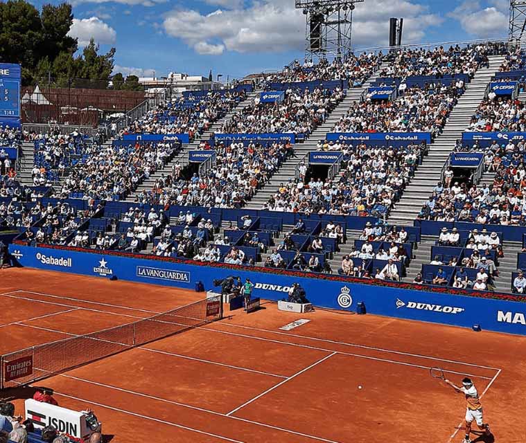 Professional tennis match on a clay court with a player serving and stadium full of spectators on a sunny day.