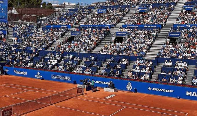 Professional tennis match on a clay court with a player serving and stadium full of spectators on a sunny day.