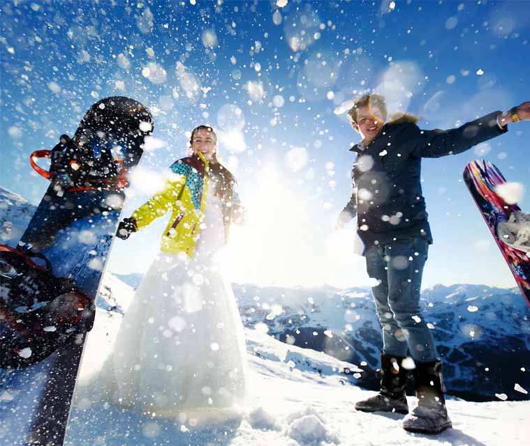 Two adults wearing colourful winter clothing, standing with snowboards in a snowy mountainous landscape under a bright blue sky, enjoying a ski holiday.