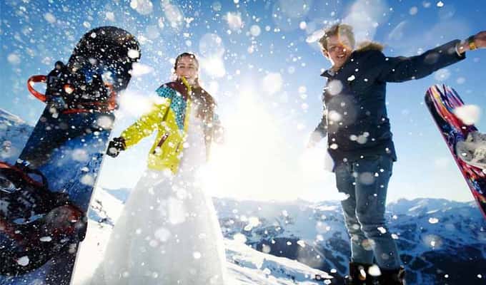 Two adults wearing colourful winter clothing, standing with snowboards in a snowy mountainous landscape under a bright blue sky, enjoying a ski holiday.