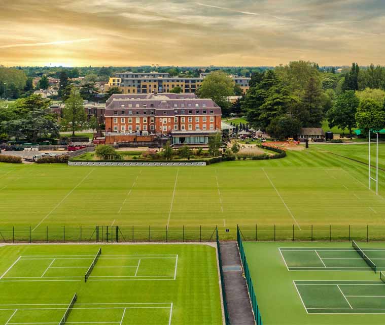A scenic aerial view of a tennis resort with grass and hard courts, and a historic hotel in the background surrounded by greenery and a sunset sky.