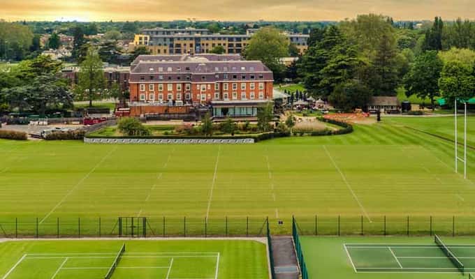 A scenic aerial view of a tennis resort with grass and hard courts, and a historic hotel in the background surrounded by greenery and a sunset sky.