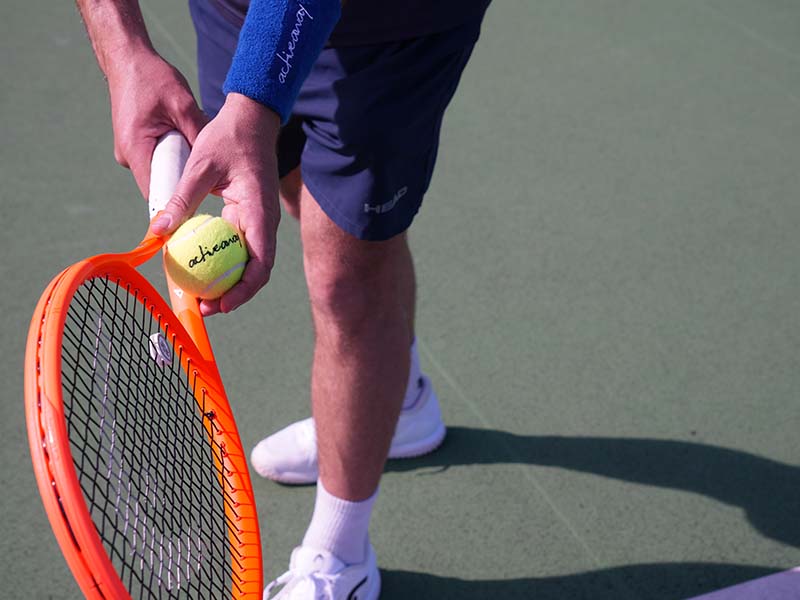 Tennis player holding a yellow tennis ball and an orange racket, ready to serve at the Active Away Junior Tennis Camps, Royal Russell School.