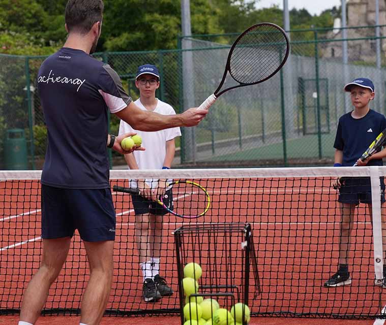 Active Away coach teaching two junior players on a clay tennis court during a junior tennis camp.