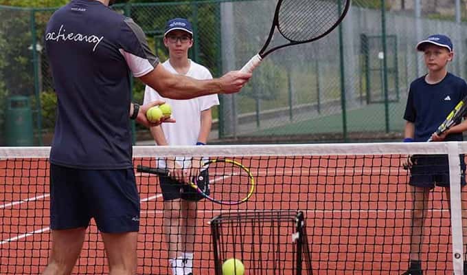 Active Away coach teaching two junior players on a clay tennis court during a junior tennis camp.