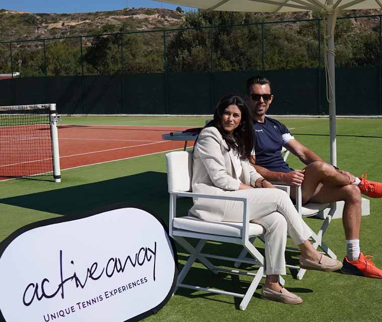 Two people seated on a tennis court beside an Active Away Unique Tennis Experiences sign, showcasing a tennis management setting.
