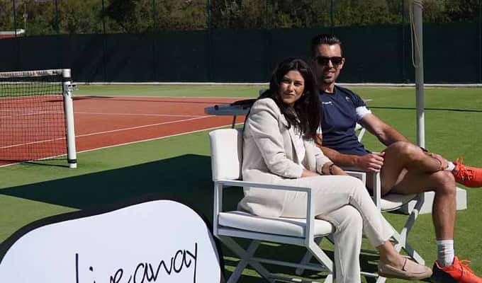 Two people seated on a tennis court beside an Active Away Unique Tennis Experiences sign, showcasing a tennis management setting.