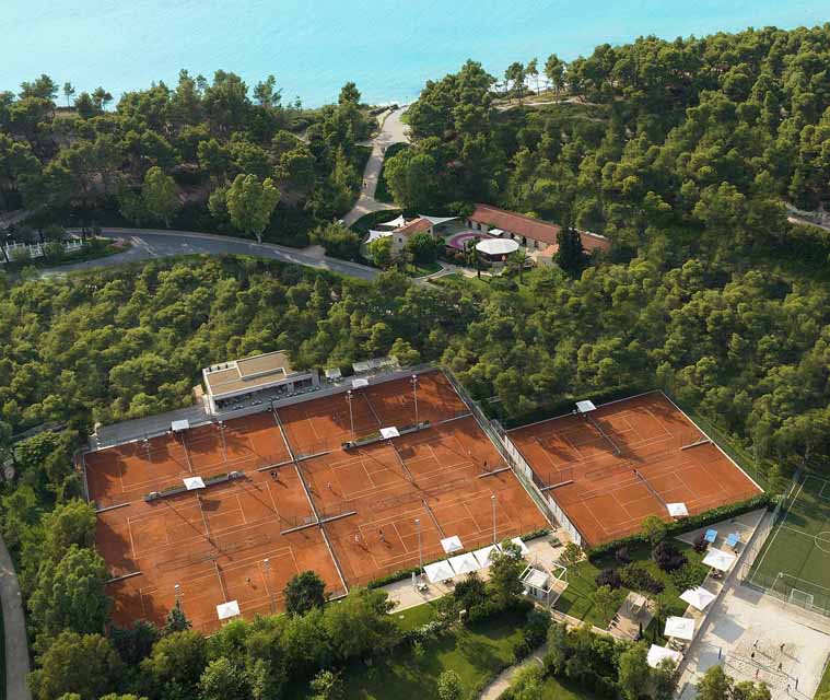 Aerial view of clay tennis courts surrounded by greenery and overlooking a body of water, ideal for tennis tours and training camps.
