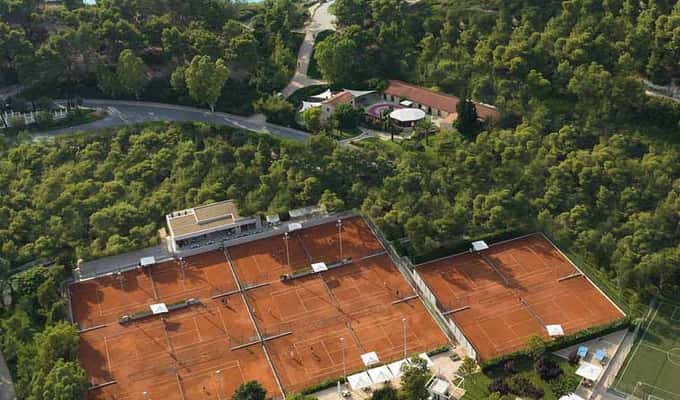 Aerial view of clay tennis courts surrounded by greenery and overlooking a body of water, ideal for tennis tours and training camps.