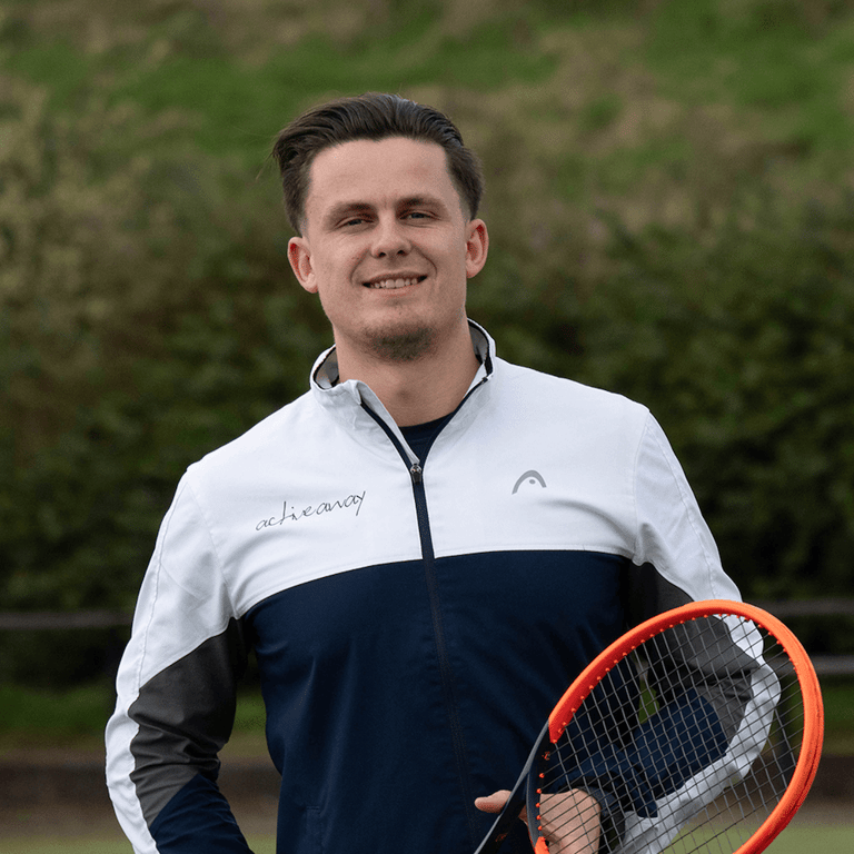 Portrait of Alex Juniper holding a tennis racket, wearing a white and navy jacket, with a backdrop of greenery.
