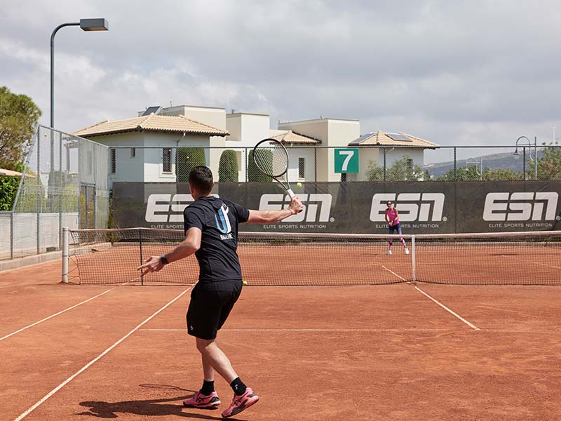 Tennis match on a clay court at Aphrodite Hills Resort with two players in action, surrounded by Mediterranean-style buildings and Elite Sports Nutrition branding.